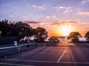 two tennis players chatting at the net with the sun setting on the sea behind them